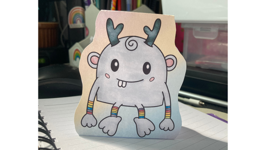A cute cartoon creature with antlers, a swirl of hair, rainbow wrists and ankles, and a big smile sits on a spiral-bound notebook. He is printed and cut to look like he is standing up on the desk.